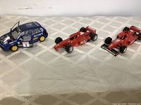 Photo showing two red Ferrari F300 Formula 1 toy cars and one blue Volkswagen Golf toy car on a white patterned surface.