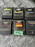 Six vintage video game cartridges on a grey cloth surface: five ColecoVision games and one Atari game. Each cartridge has a distinct label with game titles and logos. The cartridges show normal signs of age wear.