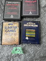 Six vintage Atari 2600 game cartridges laid out on a surface. Visible games include Space Invaders, Miniature Golf, Epyx Summer Games, and Activision Decathlon, with two additional cartridges partially visible.