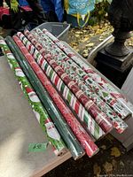 Photo of 8 rolls of various new Christmas wrapping paper laid out on a flat surface outdoors with leaves visible around.