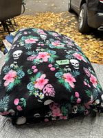 Folded black blanket featuring white skulls and colorful floral patterns on outdoor surface next to vehicles and fallen leaves.