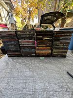 Three stacks of DVD movie cases arranged upright on a surface outdoors with trees and vehicles in background; visible titles show mostly horror and action films.