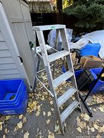 Side view of a folding aluminum ladder with four steps and visible wear, standing outdoors near a shed.