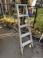 Front and side view of the aluminum folding ladder standing on pavement outdoors, showing four steps and a top platform.