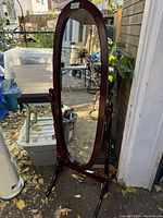 Photo of a full-length oval cheval tilting mirror with wooden frame, standing outdoors near a doorway and garden items.