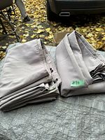 Two folded beige fabric panels laid out on a surface outdoors, showing reinforced seams and metal grommets. Possibly gazebo curtains.