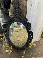 Ornate black framed wall mirror with oval mirror surface propped against a white door outside on a leaf-covered ground.