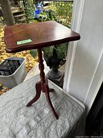 Full view of reddish vintage wooden wine table with square top and tripod base, placed outdoors on a porch.