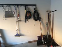 Garage wall with hanging tools and equipment including metal dust pan, mop, shovels, and brushes.