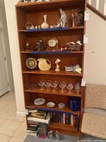 Front view of tall wooden bookcase showing five shelves with various decorative items, glassware, and books (contents not included).