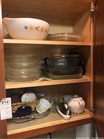 Kitchen cabinet shelves containing large Pyrex Town and Country patterned mixing bowl with floral designs, stacked clear glass bowls, rectangular glass baking dishes, ceramic teapot, small cups, and a white ceramic butter dish with lid.