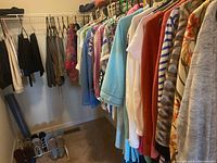 Clothing items hanging densely on a metal wire rack, including tops, sweaters, jackets, and pants in assorted colors and patterns. Shoes on floor below rack visible.