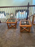 Two square wood end tables with glass tops, positioned side by side on carpeted floor beside stair railing, showing overall lot.