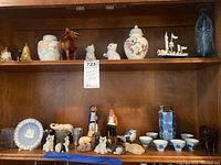 Shelf with assorted porcelain figurines, ginger jar, small bowls, shot glasses, wood animal figurines, blue glass bottle, and decorative plate.