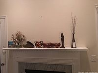 Shelf photo showing assorted decorative items including a metal copper pot, carved wooden figurine, wooden clogs, a signed pottery vase with branches, a small wooden shoe, and faux plant arrangement.