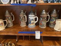 Photo showing five tall Gerz beer steins with detailed relief designs and pewter lids, arranged on a wooden shelf.