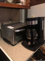 Photo showing black Hamilton Beach drip coffee maker with glass carafe and stainless steel Cuisinart toaster oven side by side on countertop.