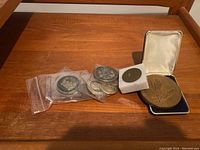 Lot arranged on two wooden shelves. Top shelf shows military medallions and coins, some in plastic sleeves, plus a large bronze U.S. Navy medallion. Lower shelf contains a pewter decanter with stopper, six small pewter cups on a tray, a black fabric-covered flask, wooden cheese board with tile insets, bird coasters, wicker basket, clear glass pitcher, and additional metal cups/flasks.