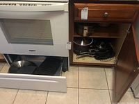 Kitchen oven with open bottom drawer containing cast iron broiling pan, pots and pans inside cabinet next to oven.