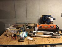 View of lot items on plywood surface against concrete wall, includes cordless drill and other tools