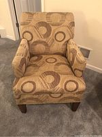 Front view of beige upholstered armchair with circular patterned fabric, padded arms, and wooden legs.