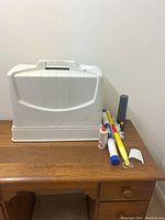 Closed white carrying case with handle on a wooden surface with small sewing accessories next to it including a tube of sewing machine oil and various small tools.