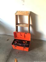 Wood step ladder next to an open orange plastic toolbox showing tools inside.