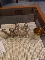 Photo showing two pairs of sterling silver salt and pepper shakers and one small sterling silver cup placed on a woven placemat on a glass table.