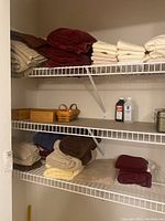 Photo showing multiple folded towels in burgundy, beige, and cream on wire shelving, with baskets and cleaning product bottles below.