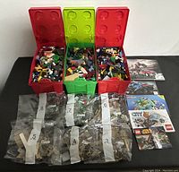 Wide view showing three large LEGO-themed plastic storage containers filled with assorted loose LEGO bricks and eight sealed numbered bags containing LEGO parts, alongside several LEGO set instruction manuals.