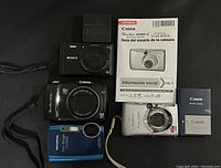 Four cameras (Sony, Canon black, Olympus blue, Canon silver) laid out with battery pack and manual visible