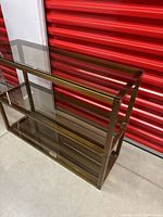 Full view of the bronze and smoked glass three-shelf shelving unit taken indoors against a red storage door background, showing the top and two lower shelves.
