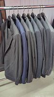 Full view of eight men's suits hanging on rack, predominantly gray with one navy blue jacket.