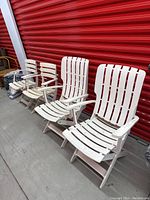 Four white foldable patio arm chairs, two large and two smaller, lined up in front of a red garage door.