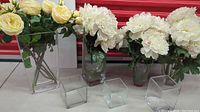 Photo showing four glass vases of different sizes holding silk flowers including white peonies and yellow roses