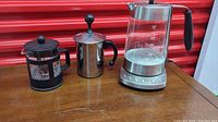 Three coffee-related appliances including two coffee presses and one electric kettle on wooden table against red background