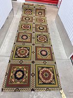 Full view of long patterned runner rug laid out on concrete floor, showing multiple decorative square blocks with floral wreath designs and geometric borders.