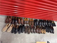 Wide view of all 21 pairs of men's shoes in front of a red backdrop showing a mix of leather and suede styles in mostly brown and black colors.