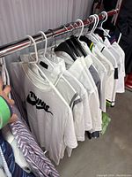 View of hangers with multiple white and one black men’s athletic sport shirts hanging on rack, showing variety of styles and brands