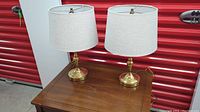 Pair of gold tone base lamps with cream linen drum shades placed on wooden table against red storage unit background