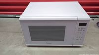 Front view of Panasonic white microwave oven with windowed door and control panel on right side, placed on concrete floor with red metal background.