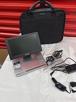 Front view of the Panasonic portable DVD/CD player with screen flipped open, power cable, car adapter, and black carrying case behind