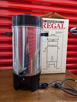 Front and side view of stainless steel Regal coffee urn with black plastic top and base along with electrical cord and spigot, placed next to original box.
