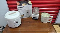 Overview of three small kitchen appliances on a wooden table against red backdrop: Aroma rice cooker with box, Cuisinart mini prep processor, Braun citrus juicer.