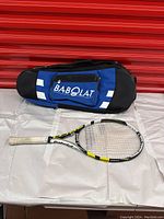 Side view of blue and black Babolat branded tennis racket bag and Babolat AeroPro Lite tennis racket with white grip on a white surface against red background.