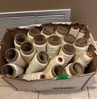 Top view of box with 22 vintage white thread cones on cardboard spools, some cones partially used, visible colored labels on some cones.