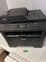 Front view of the Brother printer showing control panel, paper input/output trays, and power button.