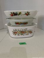 Three stacked casserole dishes including two milk glass with fruit and vegetable patterns and one clear glass with fruit pattern.