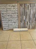 Two cork boards covered with wallpaper; one with white and gray brick pattern, the other with wooden plank pattern. Extra roll of wallpaper rolled up at bottom.