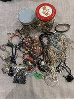 Full view of costume jewelry assortment with two jars and multiple necklaces and bracelets laid out on cloth surface